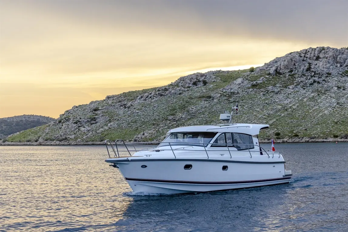 A motorboat with a white hull navigates calm waters near a rocky shore under a sunset sky.